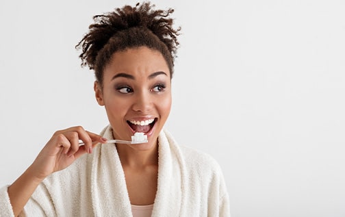 Woman with brown ponytail in fuzzy white bathrobe brushing her teeth