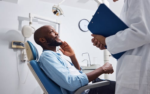 Man in blue shirt in dental chair talking to dentist with blue clipboard while touching his jaw